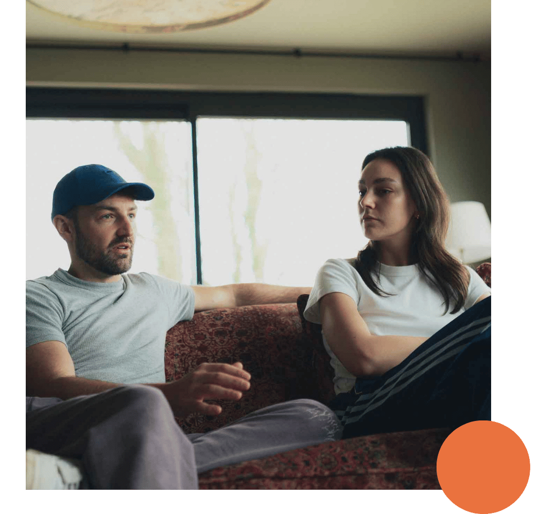 A man and woman sitting on a patterned couch in a living room, with large windows behind them. The man is wearing a blue cap and gray t-shirt, and has his arm extended along the back of the couch. The woman has long dark hair, and is wearing a white t-shirt and striped pants, looking at the man with a neutral expression.