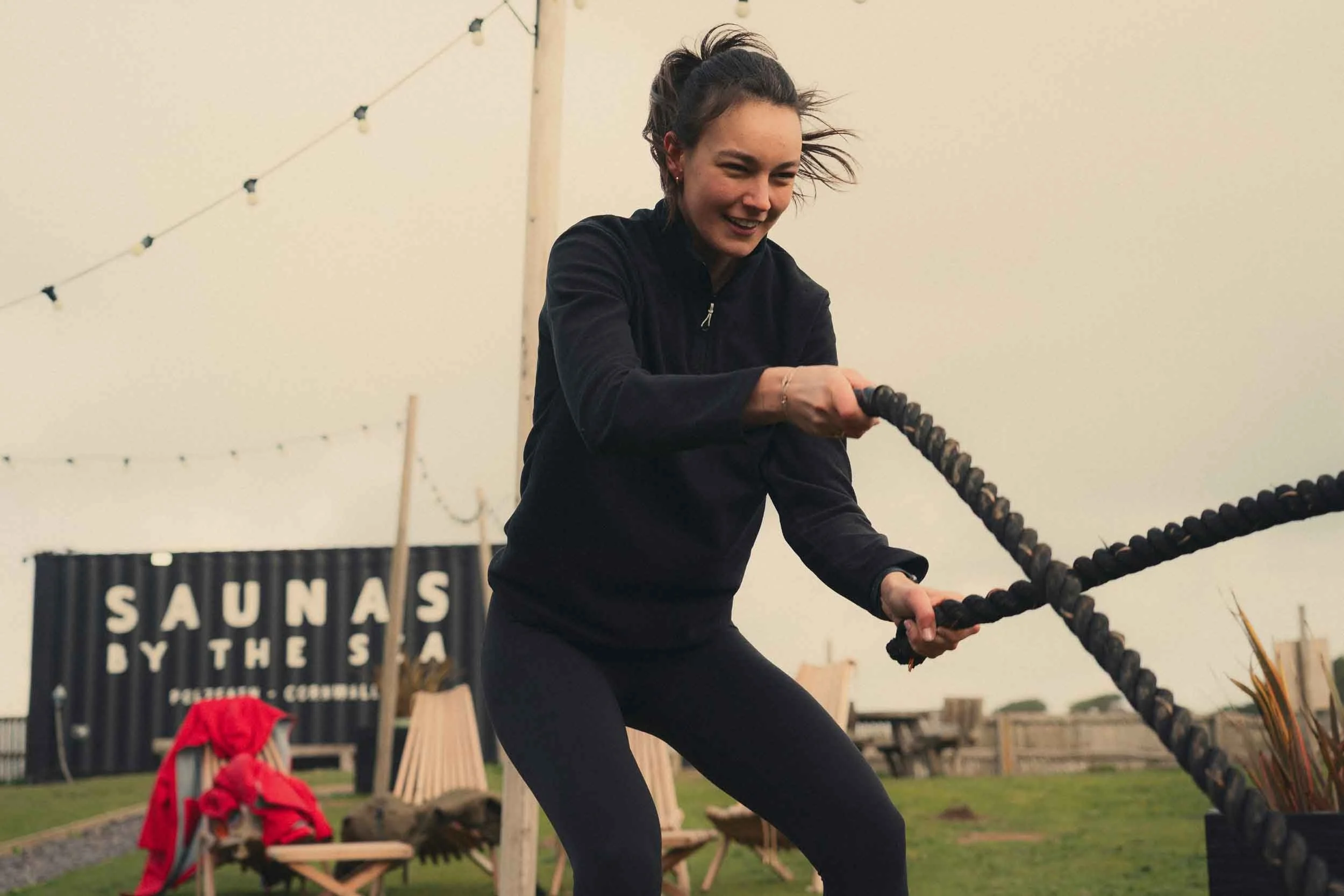 A woman with curly hair tied back, dressed in black, is engaged in a workout with battle ropes outdoors. She is smiling, and wind is blowing her hair. In the background, there is a sign that reads "SAUNAS IN THE S.