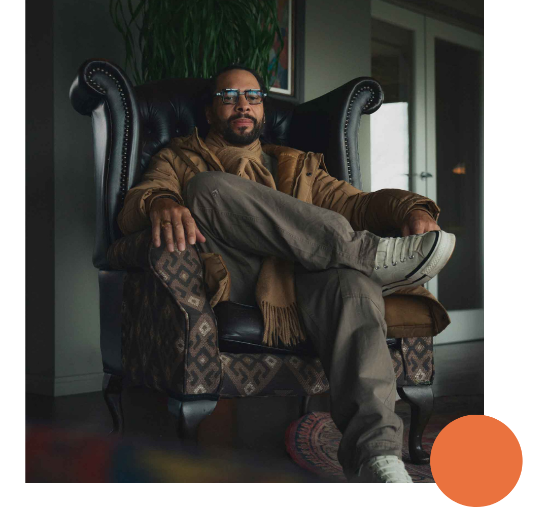 A man with glasses and a beard sitting in a large black armchair with wooden and fabric details, wearing casual clothing and a scarf, in an indoor setting.