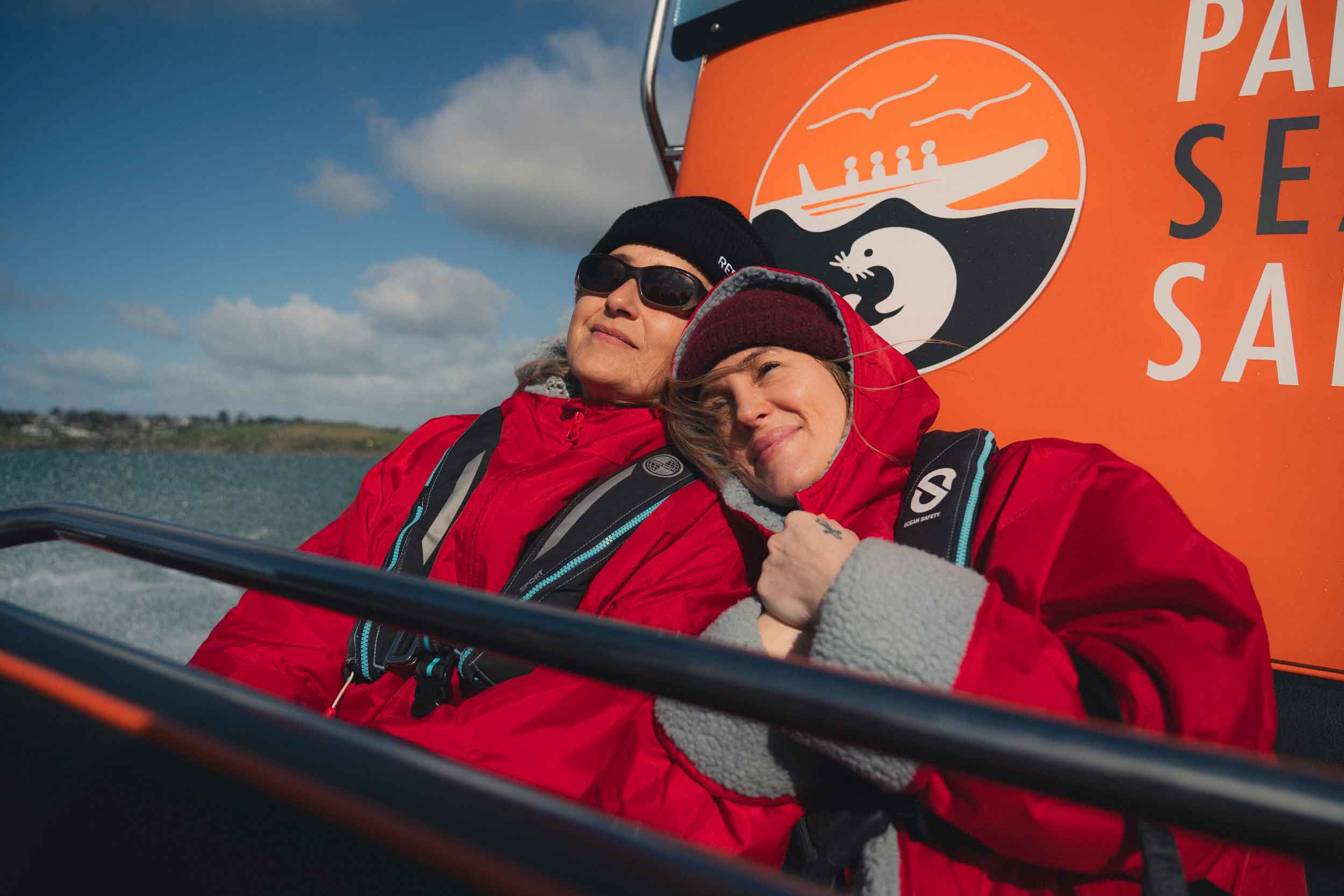 Two people dressed warmly in red jackets, one wearing sunglasses and a black beanie, are taking a selfie in front of a bright orange sign with a whale and a sunrise logo, near a body of water and a partly cloudy sky.