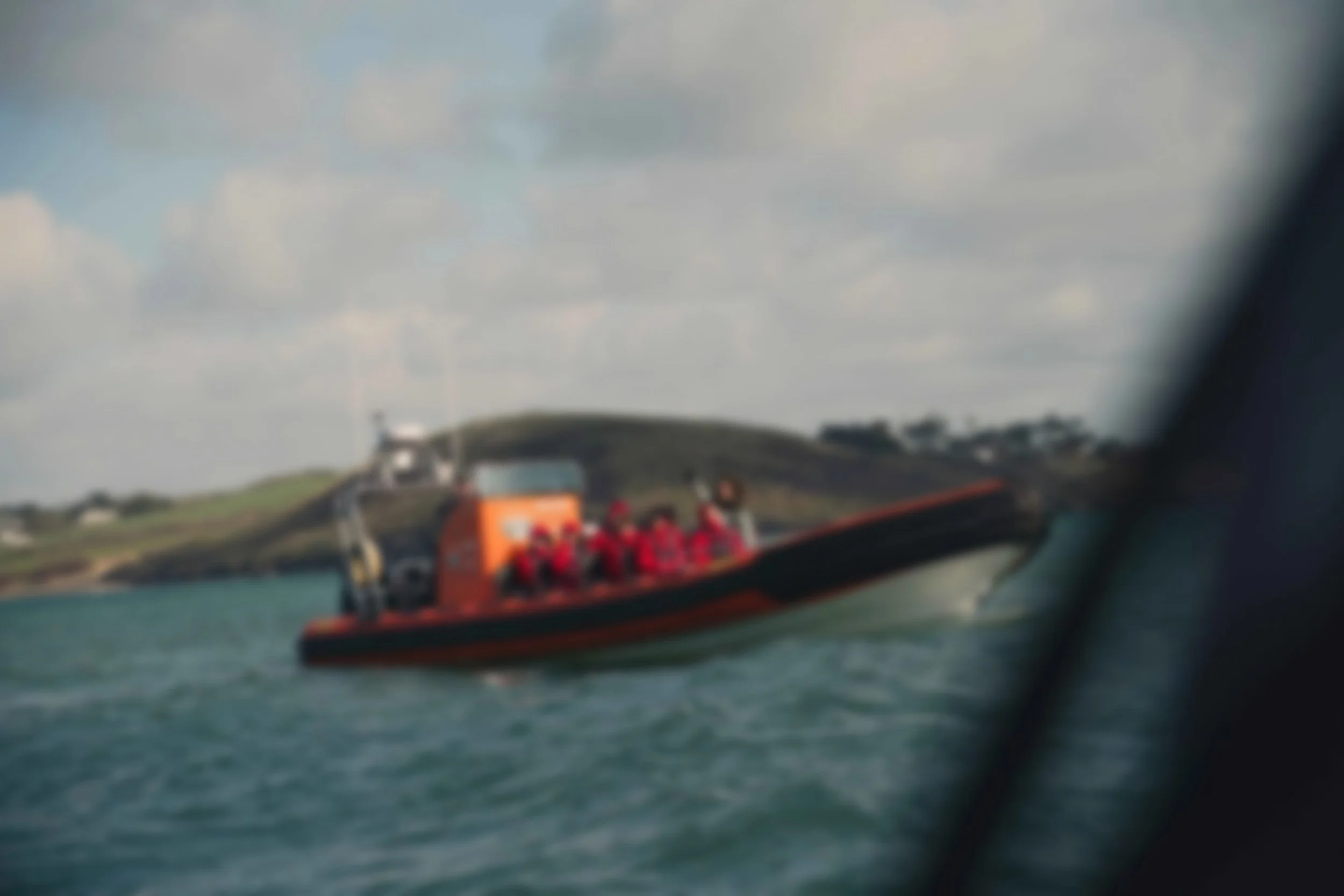 A boat with passengers wearing red life jackets on water, with hills and cloudy sky in the background.