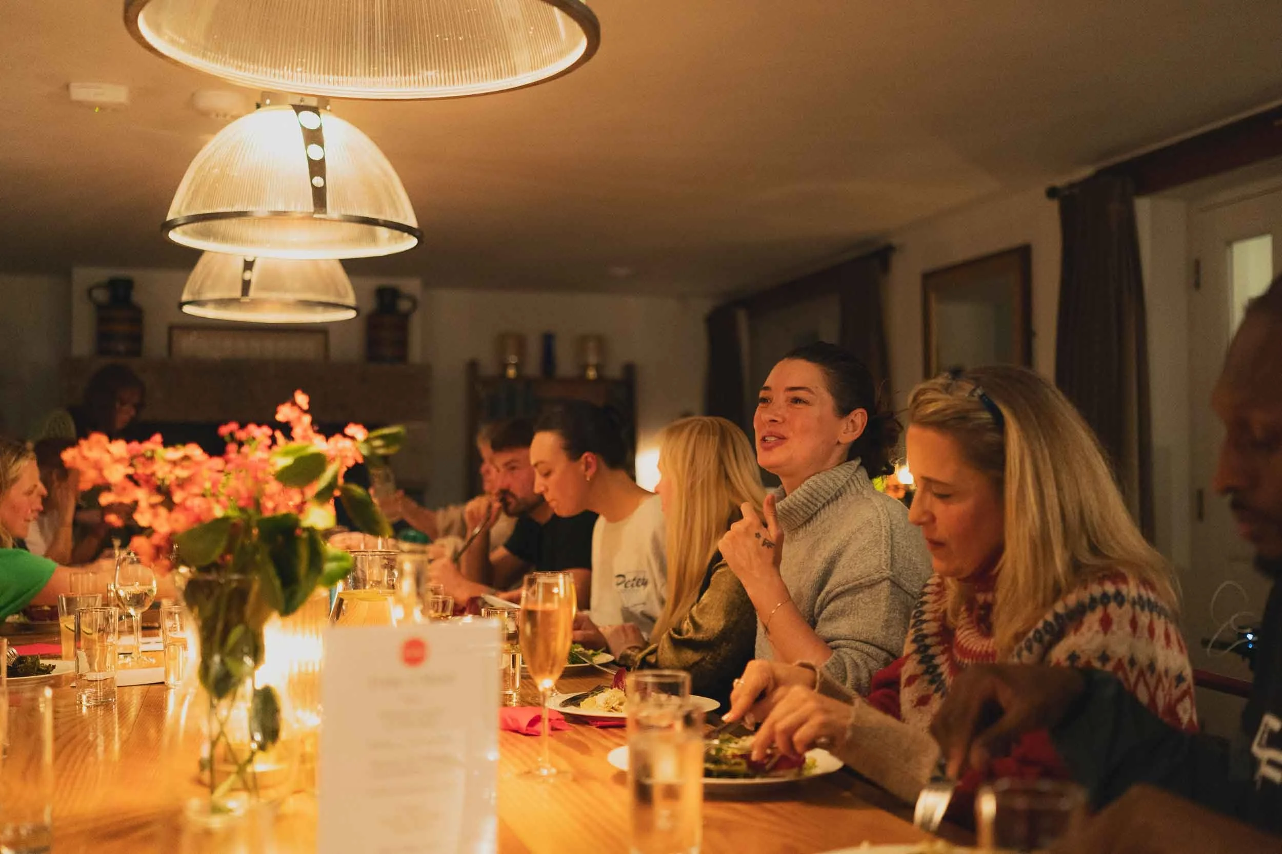 People dining at a long table in a warmly lit room with modern overhead lights and a colorful floral centerpiece.