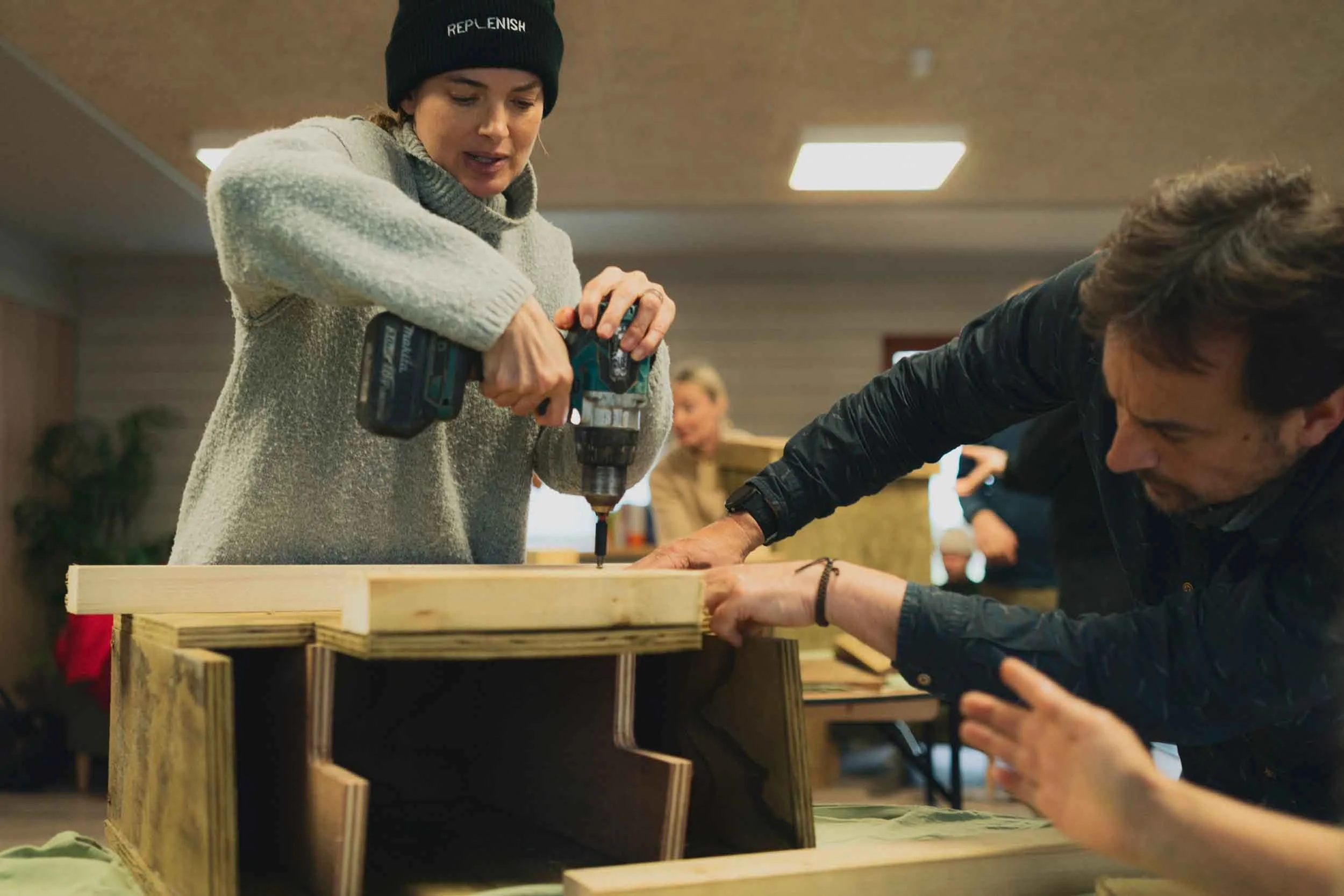 A woman wearing a black beanie and gray sweater is using a cordless drill to assemble a wooden furniture piece inside a room with wooden walls and ceiling. A man in a black jacket is holding the furniture steady while sitting at the same table. In the background, besides the people, other furniture or objects are visible.