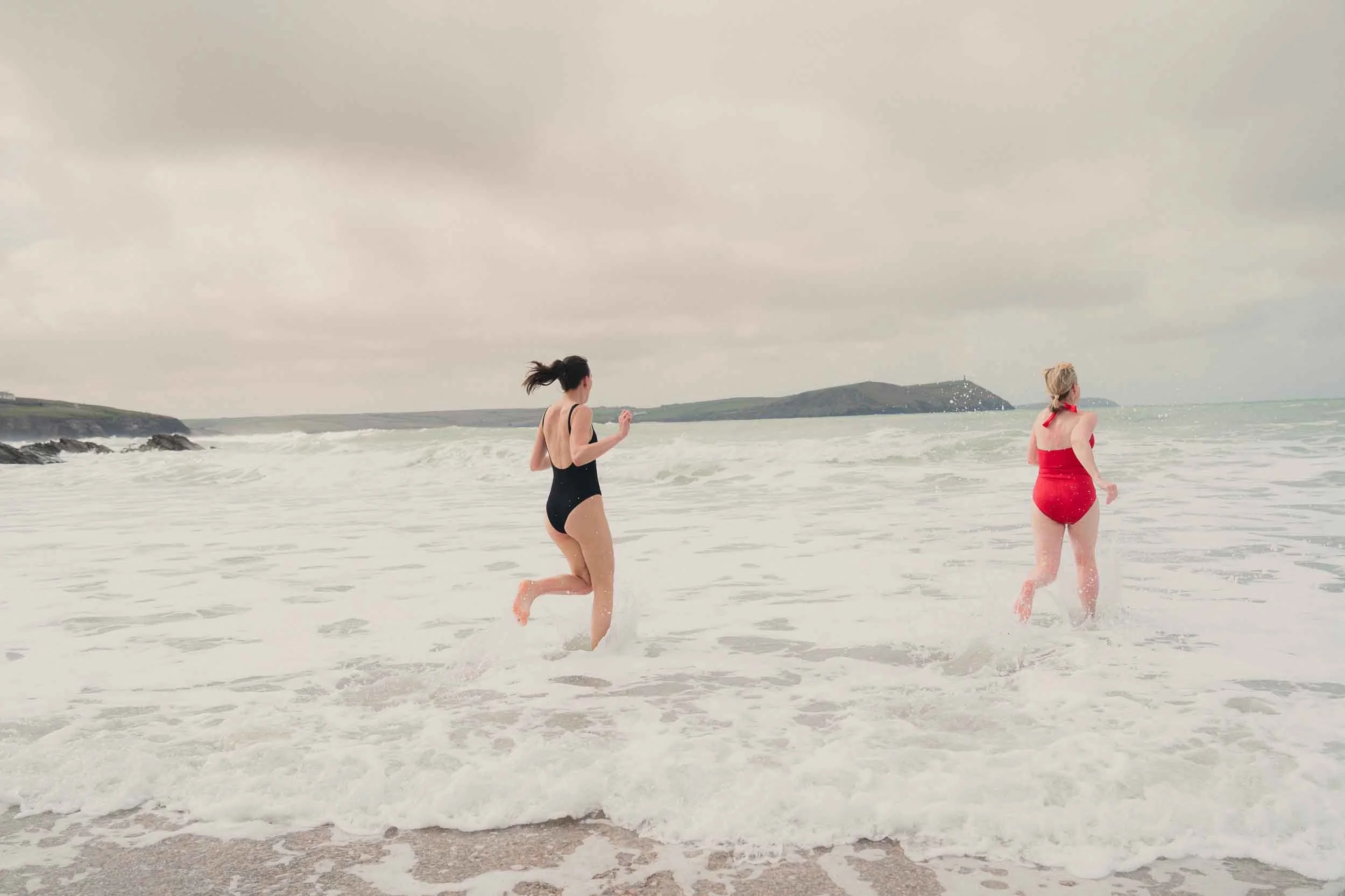 Two women in swimsuits running out of the ocean on a cloudy day, with an island and rocky coastline in the background.