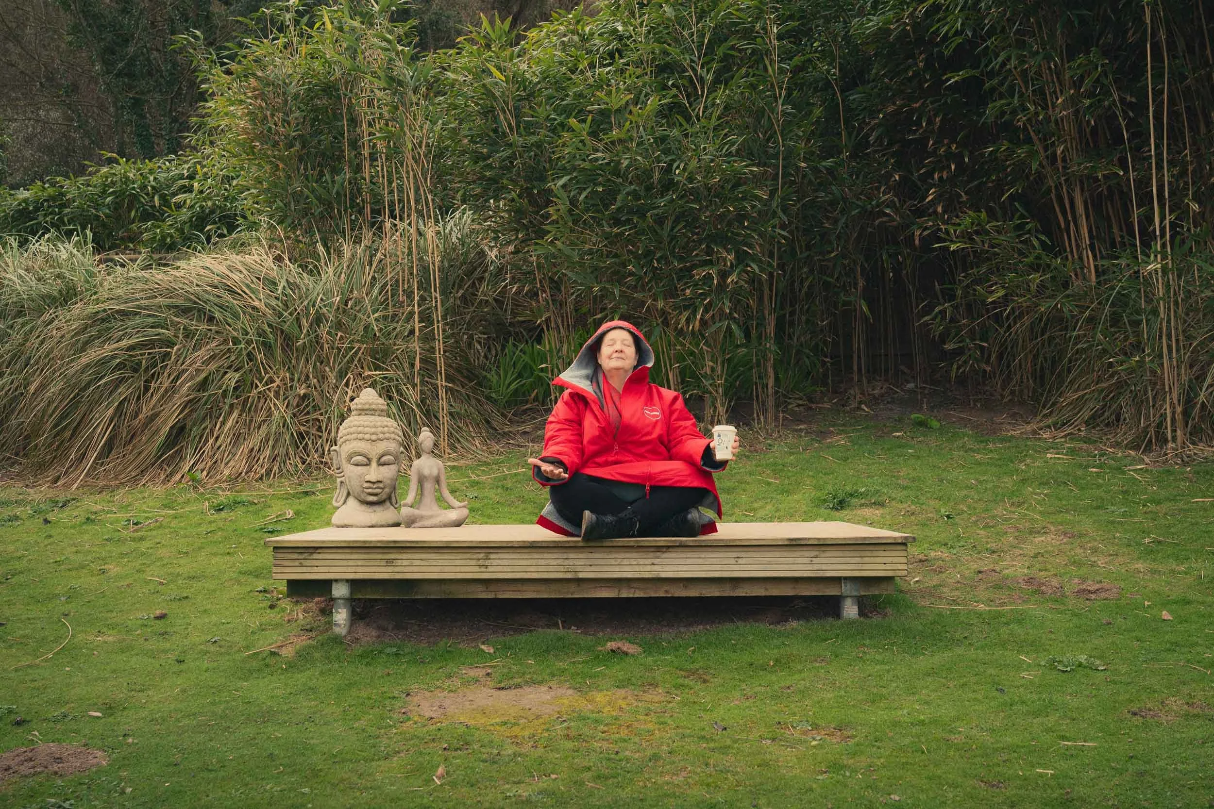 A woman meditating outdoors in front of dense green foliage, sitting cross-legged on a wooden platform with stone statues, wearing a red jacket with a hood, holding a cup in one hand and a phone in the other.