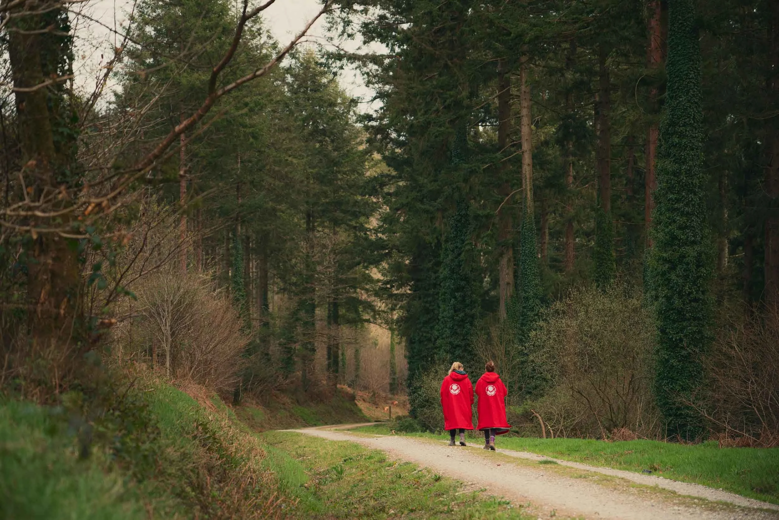 Two children wearing red jackets walking on a dirt path through a forest with tall trees and green foliage.