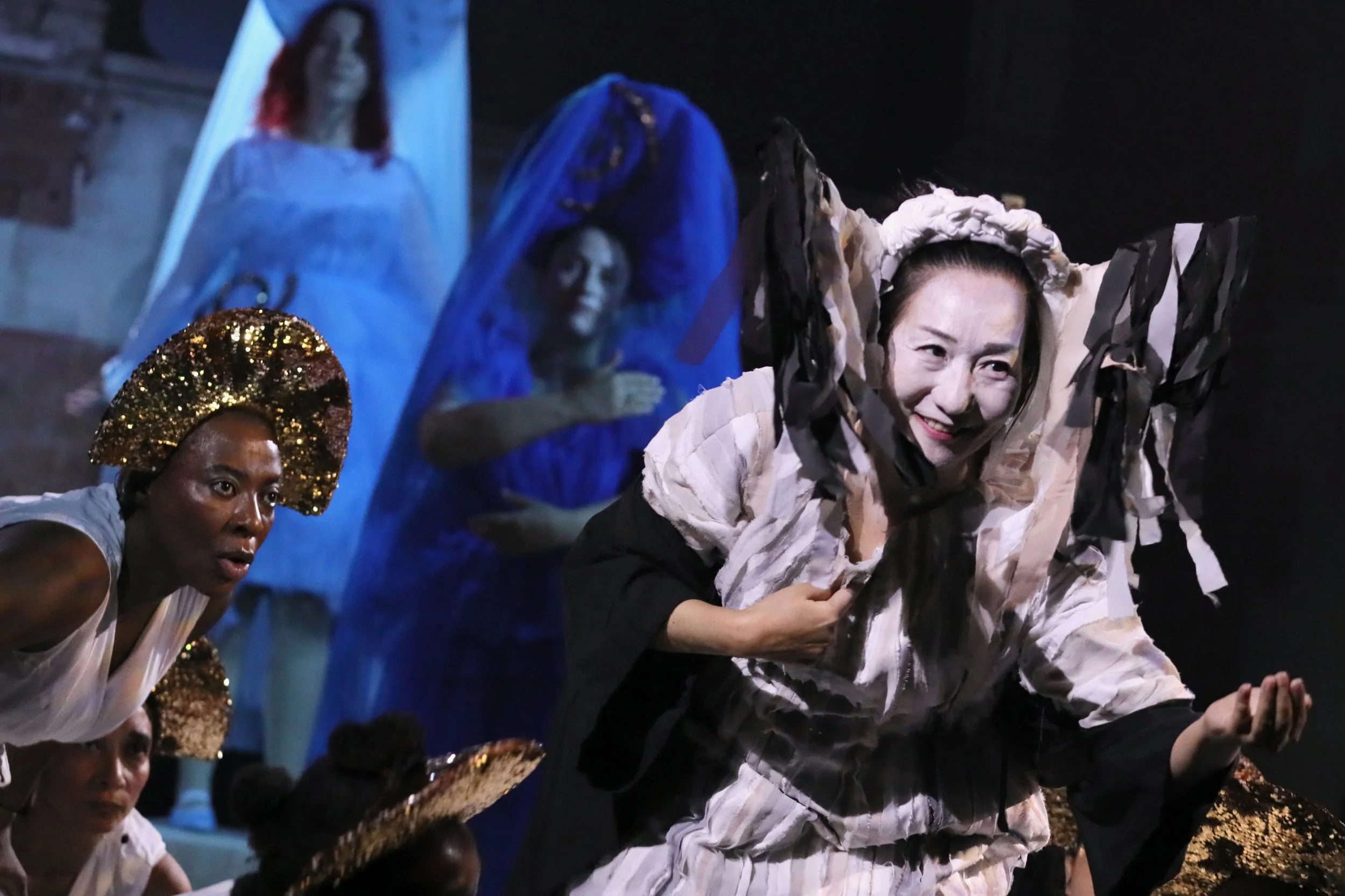 Chisato Minamimura in fringed headpiece and costume playing self loathing other cast in the background.