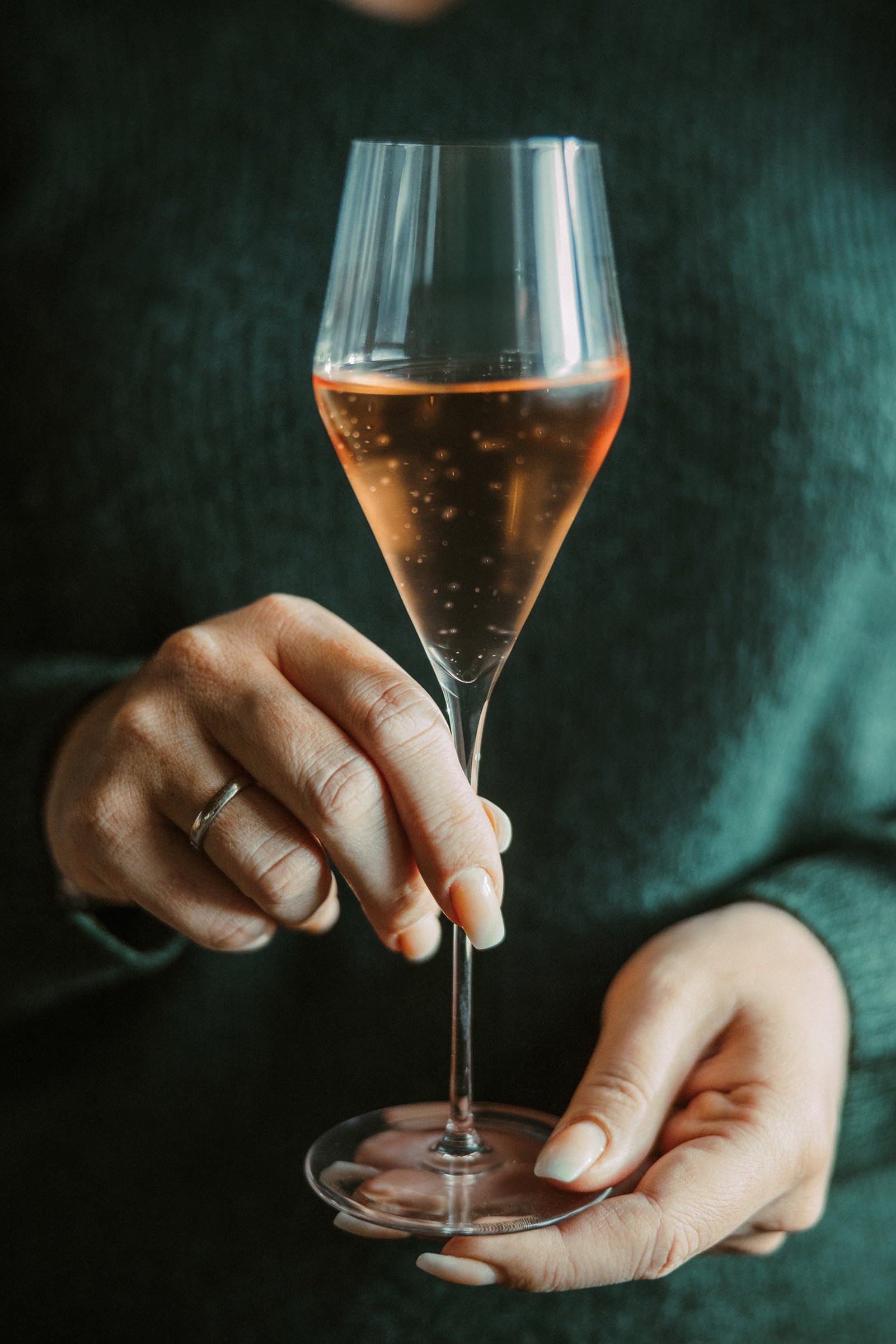 Eine Frau hält ein Champagnerglas, gefüllt mit Frizzante Rosé, vor ihrem Oberkörper. Sie trägt einen dunkelgrünen Pullover, an der Hand einen Ring am Finger.
