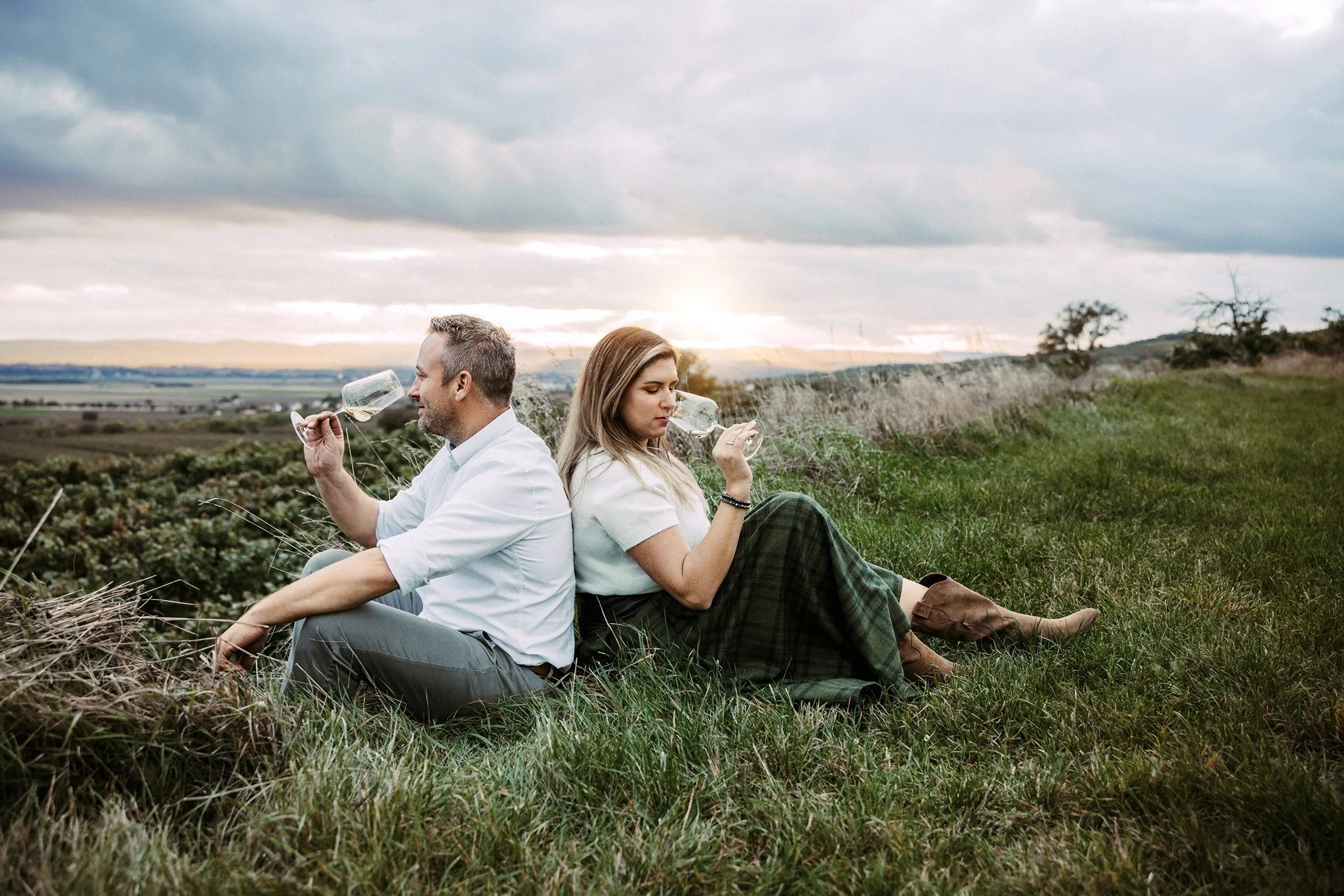 Gerald und Theresa Waltner sitzen Rücken an Rücken im Feld und trinken ein Glas Wein während des Sonnenuntergangs am Wagram.