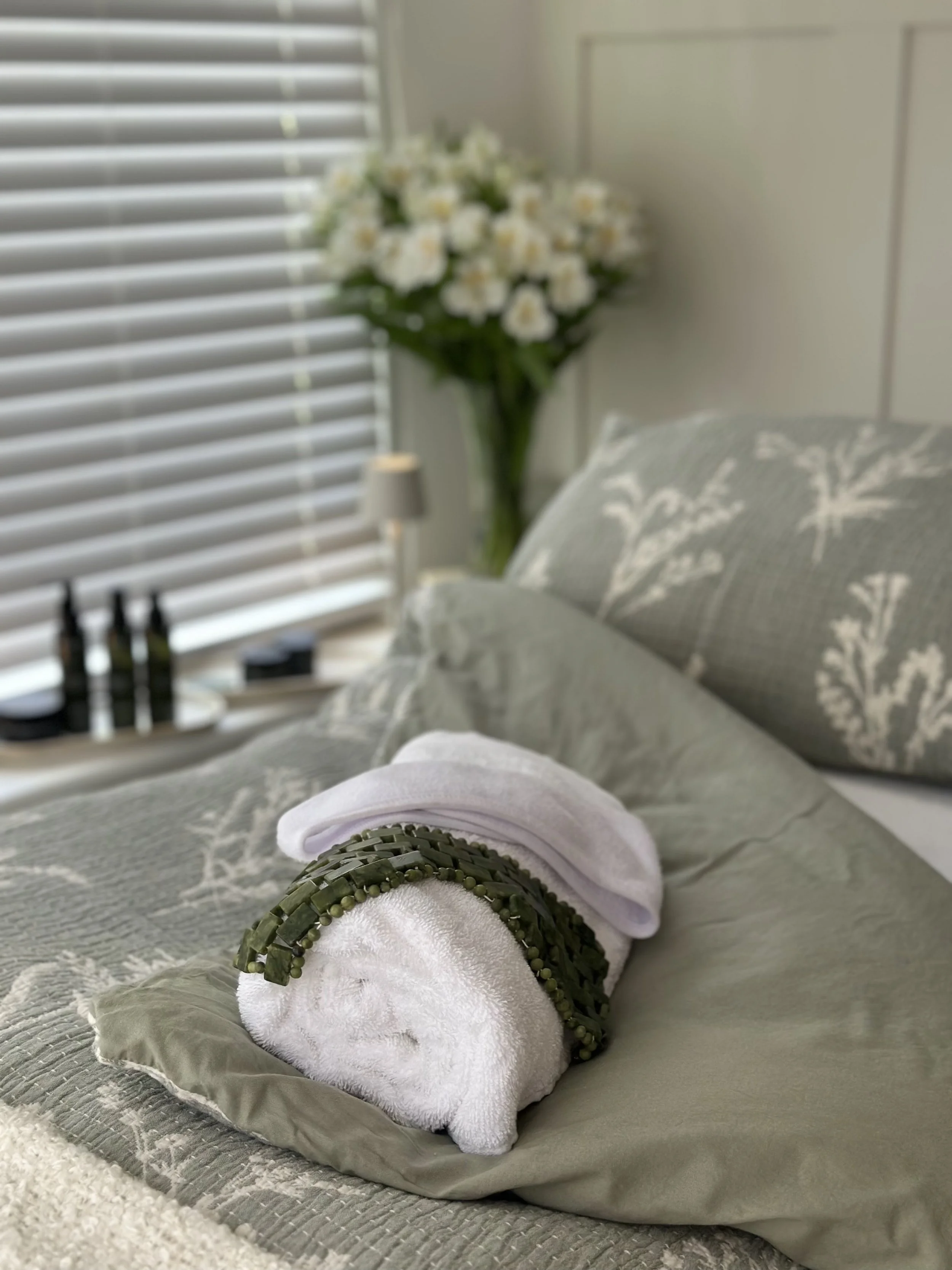 A facial treatment scene with a bed, a rolled white towel with a green beaded eye-mask, a green pillow with white pattern, a floral arrangement, and window blinds in the background and facial skin care products arranged on a shelf.