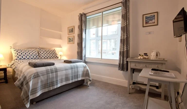 A cozy bedroom with a window, bed with patterned pillows and a plaid blanket, bedside lamps, wall art, and a small desk with a TV and kettle.