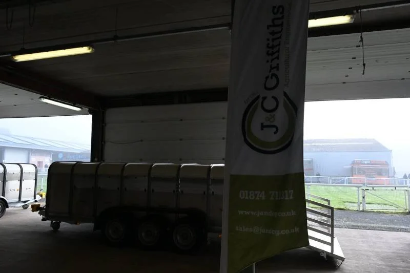 Inside a warehouse or storage shed with a large trailer or container and a vertical flag with the logo 'J&C Crichts' and contact information. The open garage door reveals storage and a grassy outdoor area.