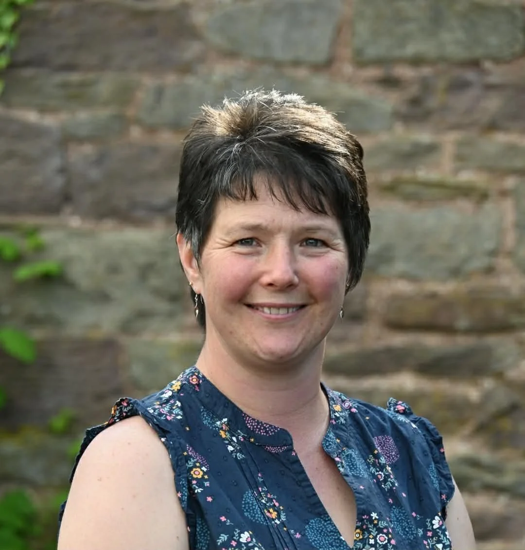A woman with short dark hair, smiling, wearing a sleeveless blue floral top, standing outdoors in front of a stone wall with green leaves.