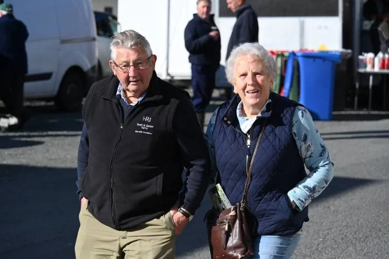 An elderly man and woman walking outdoors, smiling, in a parking lot with vehicles and a food stand in the background.