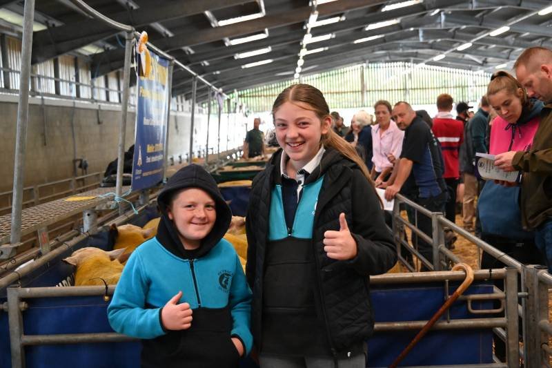 Two children at an indoor farm event, smiling and giving thumbs up, with a crowd of people and animals in pens behind them.