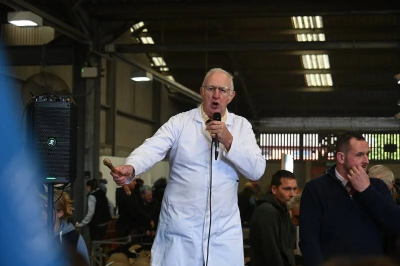 Man in a white lab coat holding a microphone and a sausage, speaking to an audience in an indoor setting.