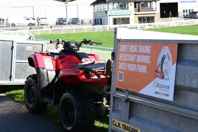 Red all-terrain vehicle (ATV) parked next to a decorative farm-themed sign warning about resistant worms, with a grassy field, pathway, and buildings in the background.