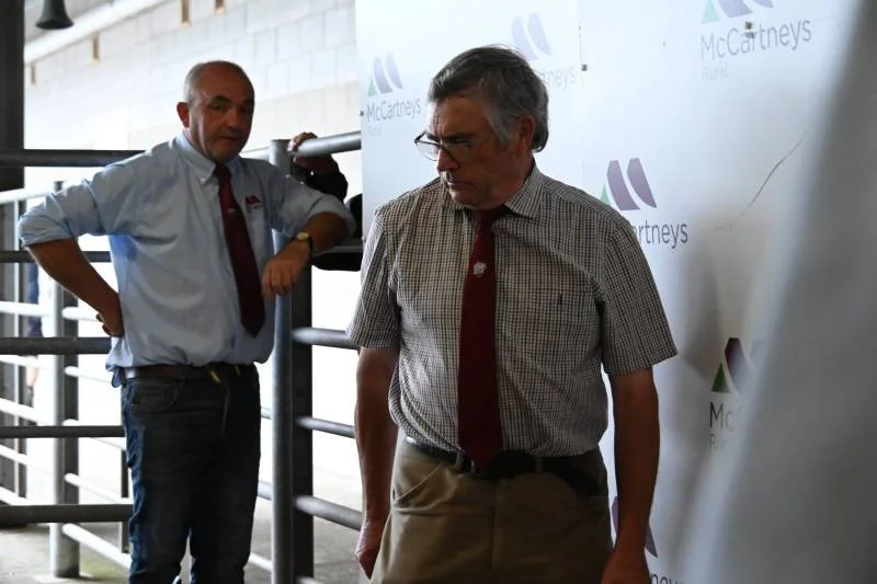 Two men standing indoors near a railing, one looking down and the other looking at him, with a McCartneys banner in the background.
