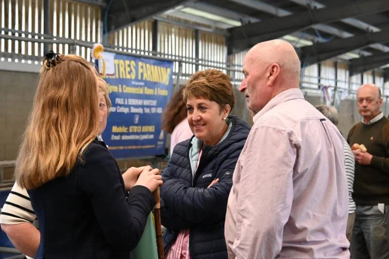 A group of people engaged in conversation at an indoor event, with a woman with red hair and a black jacket talking to a man with a bald head and light-colored shirt, while another woman with short brown hair and a dark jacket smiles nearby.