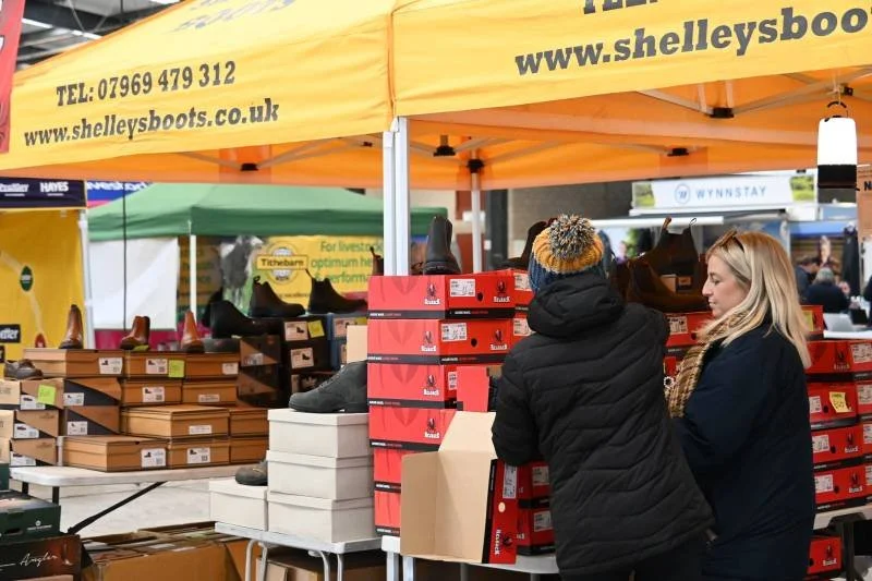 Two women shopping for shoes at an outdoor market, with stacks of shoe boxes and a yellow tent overhead.