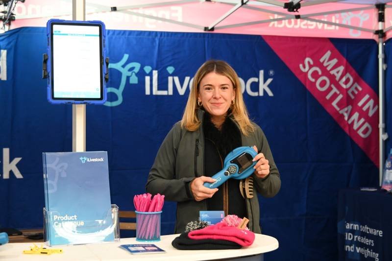 A woman standing at a booth with a blue backdrop that displays 'Livestock' and 'Made in Scotland.' She is holding a blue handheld electronic device and smiling, with pink pens and a pink cloth on the table in front of her.