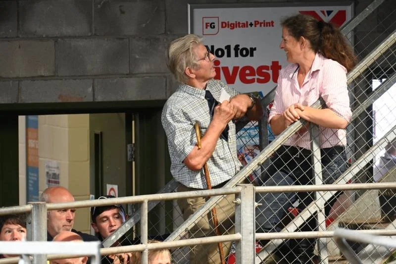 An elderly man with gray hair and glasses, holding a walking cane, is greeting a young woman with curly hair on a staircase, both smiling and shaking hands. Several people are seated nearby, and there's a sign in the background for a print and digita
