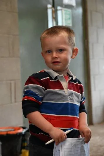 Young boy with short blond hair wearing a striped long-sleeve polo shirt, standing indoors holding a paper and a pen.