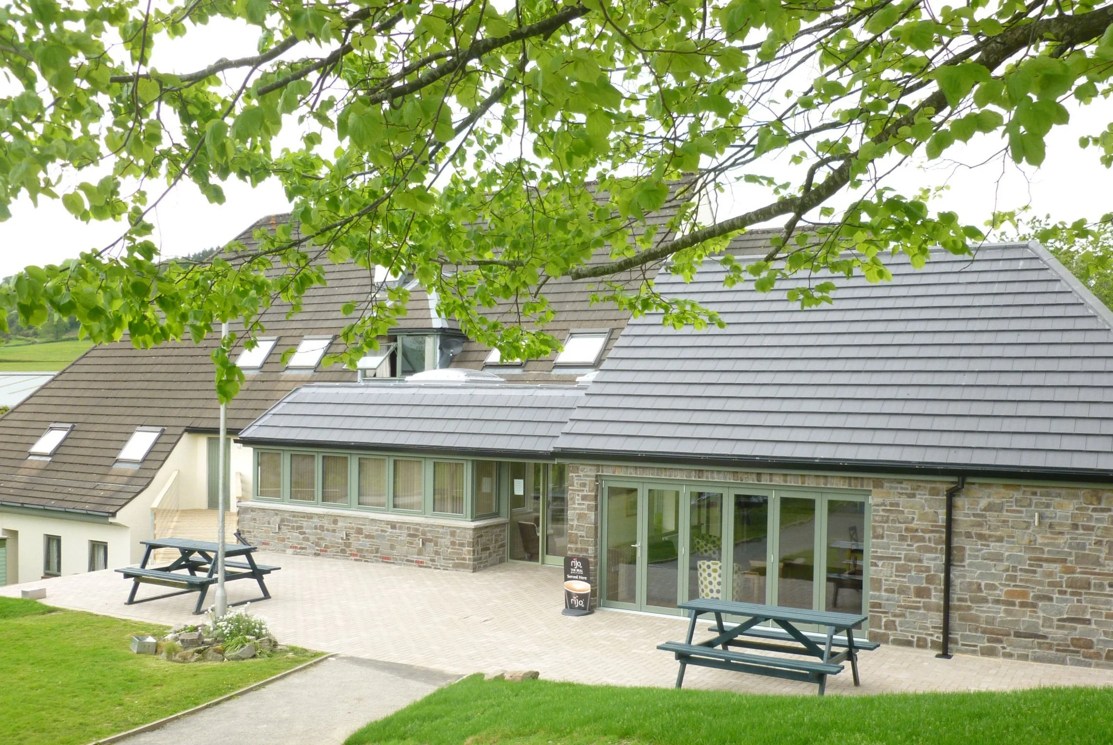 A building with a sloped roof partially obscured by green tree branches with leaves, a patio with benches, and a well-maintained lawn in the foreground.