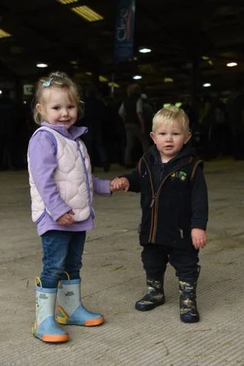 A young girl and boy holding hands at an indoor event. The girl is wearing a purple jacket, blue jeans, and rain boots, and is smiling. The boy is dressed in a navy jacket, dark pants, and rain boots, and has a neutral expression.