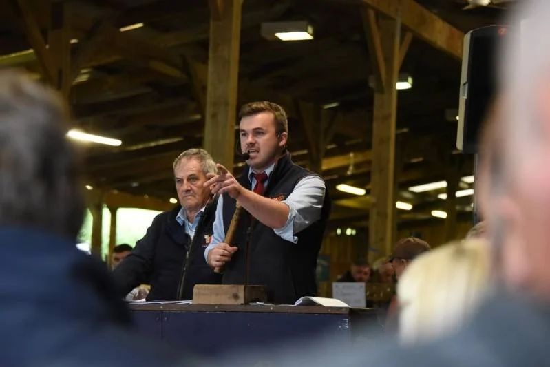 Man speaking at a gathering in a wooden barn, with an older man standing beside him, audience in the foreground.