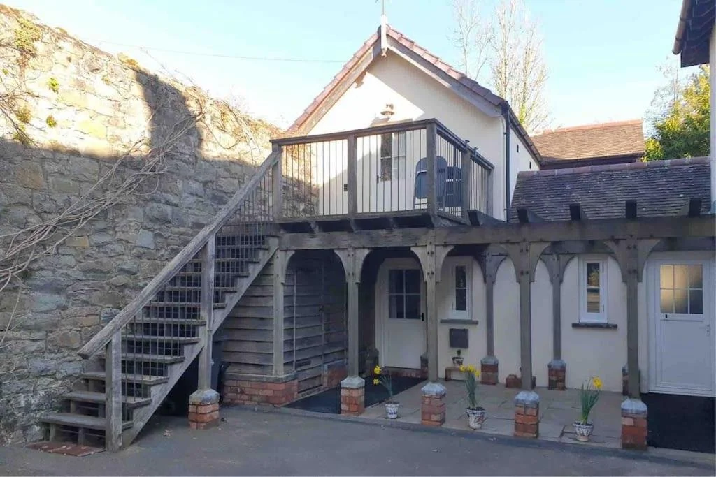 View of the exterior of a house with an upper terrace accessed by stairs, supported by wooden beams and brick pillars, with potted flowers on the ground level.