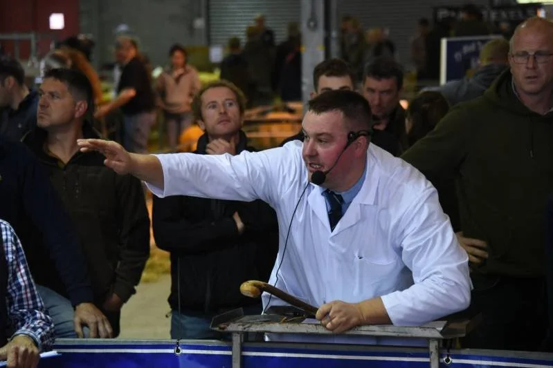 A man in a white jacket with a headset and microphone is speaking passionately, gesturing with his left arm extended, during a public auction or demonstration. Several people are watching him, some with arms crossed, and others looking attentive, in 