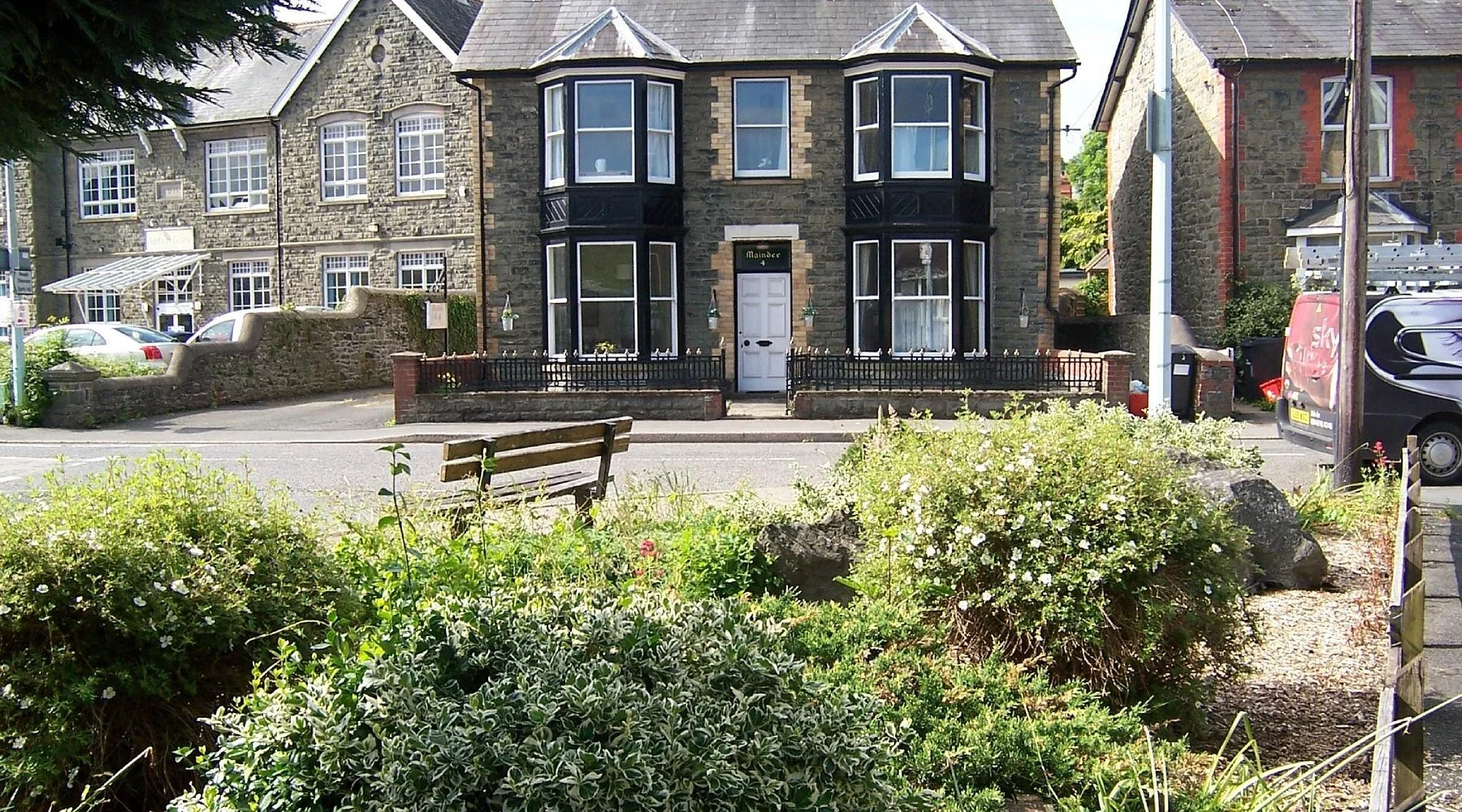 A stone house with a white front door, black window frames, and bay windows on a sunny day. There is a small fenced garden area in front, a bench, and plants along the sidewalk. The street has parked cars, and a car wash sign is visible on the right.