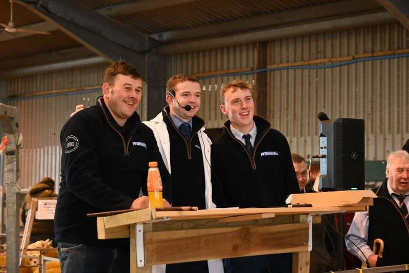 Three young men standing behind a wooden counter, smiling at the camera, in an industrial or warehouse setting. One is wearing a black jacket, another a white jacket with a headset, and the third a black jacket with a zipper. There is a computer moni