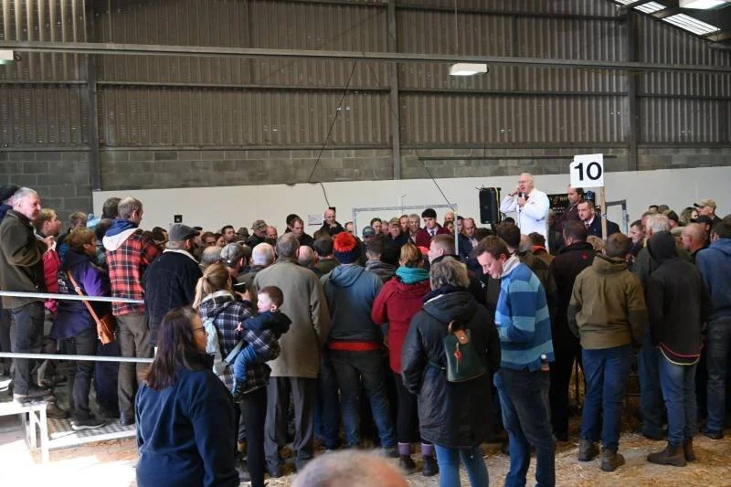 A speaker standing on a raised platform with a microphone addressing a large crowd gathered inside a warehouse or industrial building, with a numbered sign '10' nearby.