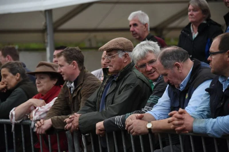 A group of people standing behind a metal barrier at an outdoor event, some wearing jackets, hats, and glasses, with a tent in the background.