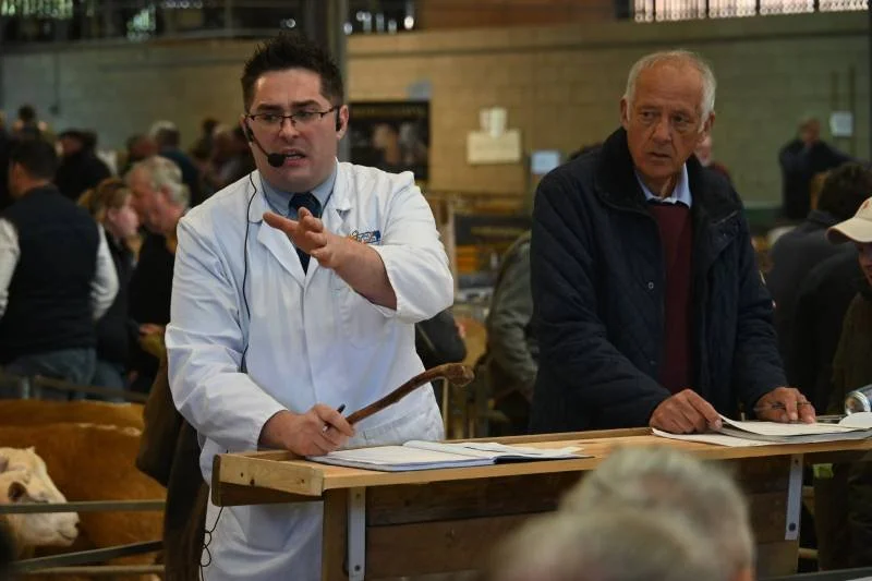 A man in a white lab coat and glasses is speaking, holding an object, with a crowd in the background, at what appears to be an auction or auction-like event.