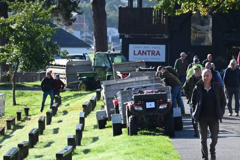People walking near parked utility vehicles and a sign advertising 'LANTRA' in an outdoor setting with trees and a grassy area.