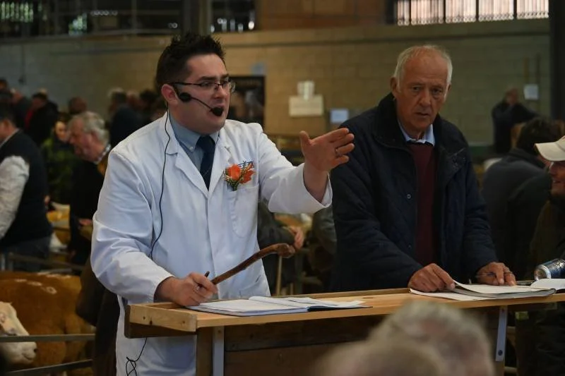 A young man wearing glasses, a white lab coat with a flower pinned to it, and a headset is speaking and gesturing with his hand at a wooden counter. An older man in a dark jacket stands next to him, listening. They are in a busy indoor setting with m
