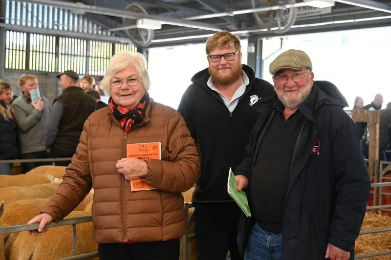 Three people visiting a livestock show, with sheep in pens behind them. The woman on the left is wearing glasses, a brown jacket, and a scarf, holding a booklet. The man in the middle has glasses, a beard, and is wearing a black jacket. The man on th