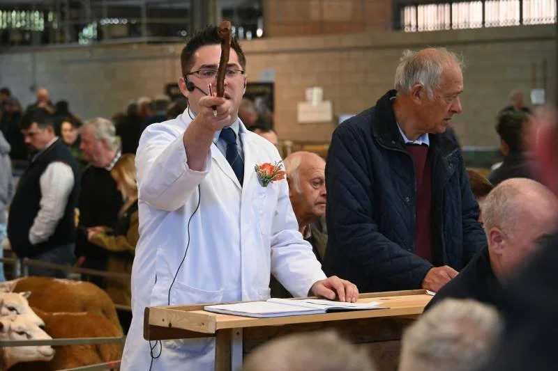 Man in white lab coat holding a pen or indicator, standing at a wooden podium meditating during an indoor event with a large crowd.