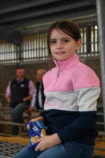 Young girl in pink, gray, and navy jacket sitting on a bench, holding a snack, with people in the background.