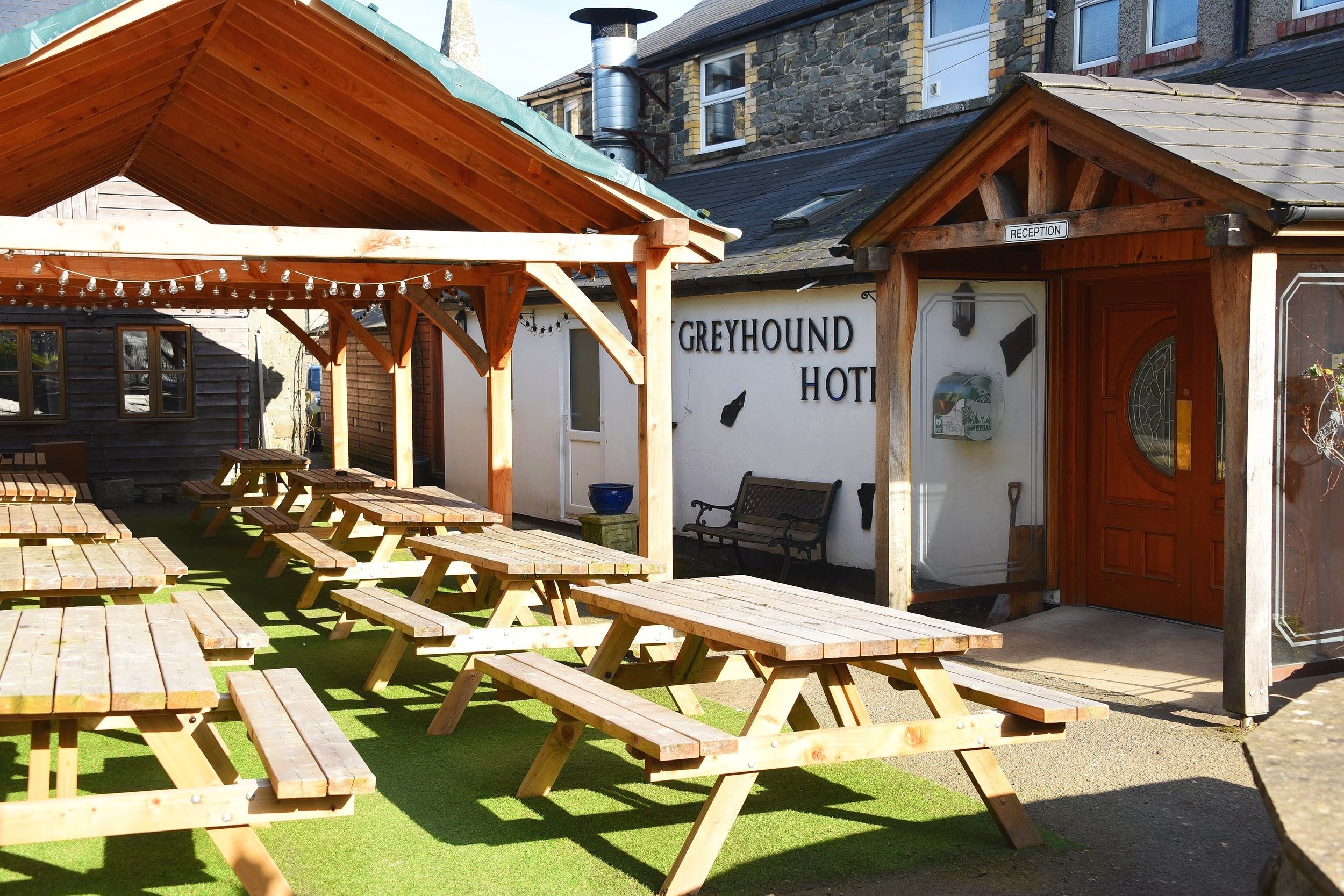 An outdoor patio area with wooden picnic tables and benches, string lights overhead, adjacent to a building with a wooden pergola labeled 'Reception' and a sign for 'Greyhound Hotel' on the wall.