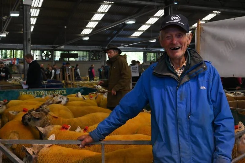 An elderly man in a blue jacket and black cap smiling at a livestock auction, standing behind a pen of yellow sheep. Other people and livestock are visible in the background inside a large indoor barn.