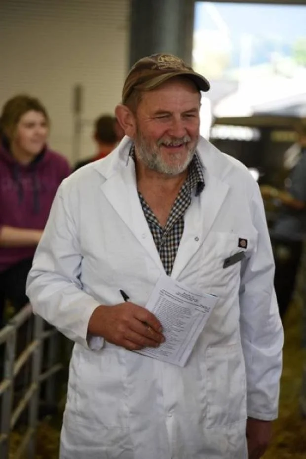 A man in a white lab coat holding a clipboard, smiling, in what appears to be an indoor farm or agricultural setting with a woman in the background.