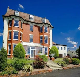 Multi-story brick building with large windows, surrounded by greenery, with a small porch and a pathway leading to the entrance.