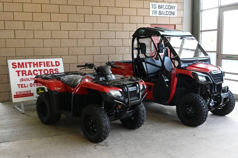 Two red utility vehicles, a quad and a side-by-side, parked indoors near a brick wall. There is a sign for Smithfield Tractors and a sign pointing to the balcony.