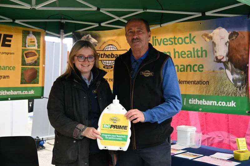 Two people standing under a green canopy at a livestock event, holding a bottle of livestock supplement, with banners about livestock health and performance in the background.