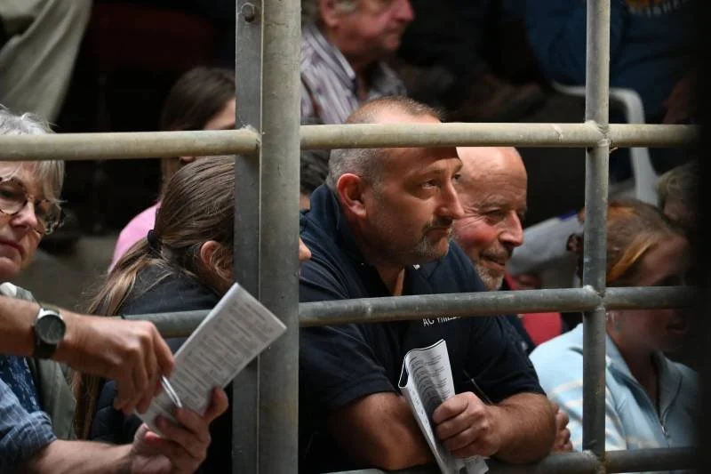 People sitting behind and around a metal barrier, watching an event, some holding papers or programs, with attentive expressions.