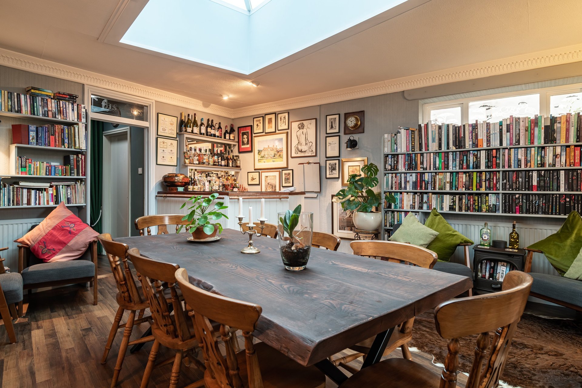 A cozy dining area with a wooden table, surrounded by wooden chairs adorned with cushions. The room features bookshelves filled with books, framed artwork on the walls, a potted plant, and a skylight allowing natural light to brighten the space.